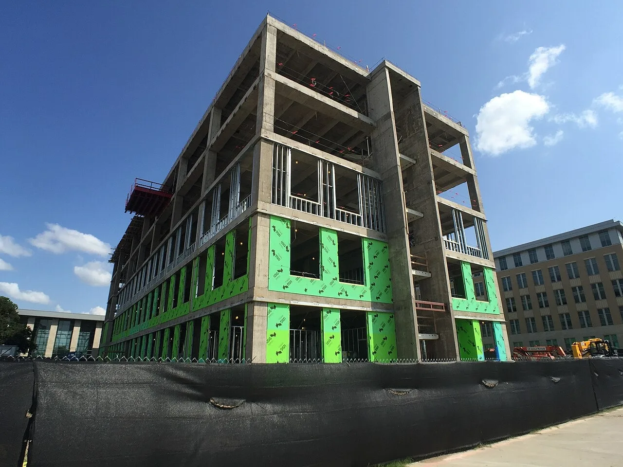 A multi-story concrete building frame going up behind a black site fence in the Agriculture and Life Sciences Complex at Texas A&M University, with an orange lift and a yellow machine at the right edge