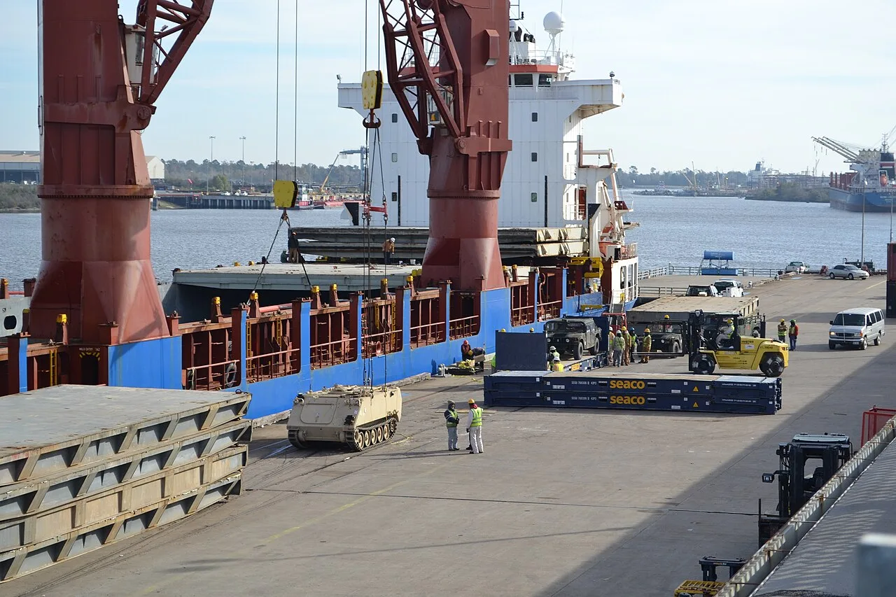 A cargo ship using its own deck cranes at a Southeast Texas dock, with forklifts and stacked cargo on the wharf, during an Army cargo operation at the ports of Beaumont and Port Arthur