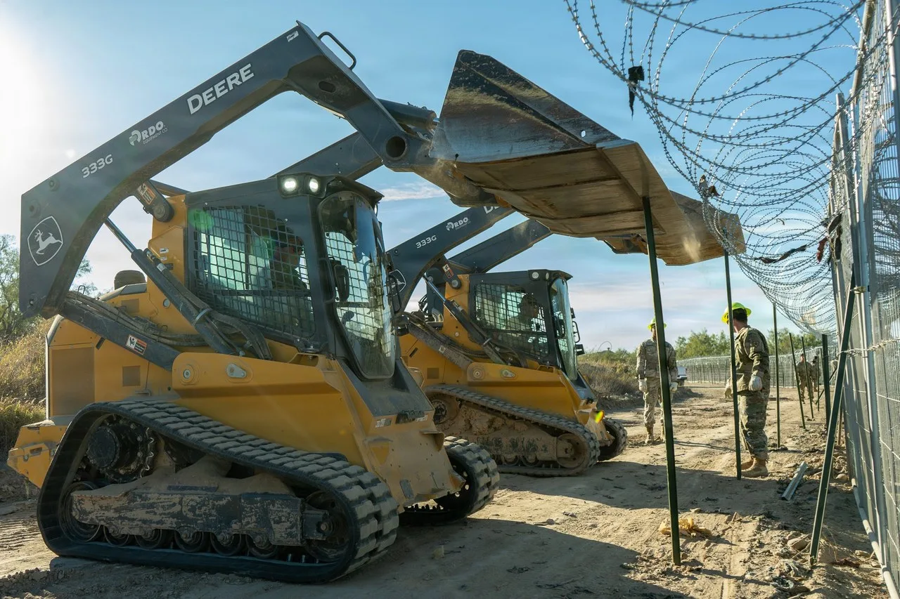 Two yellow John Deere 333G compact track loaders parked side by side with their arms raised beside a wire-topped fence near Eagle Pass, Texas, with soldiers in camouflage standing by