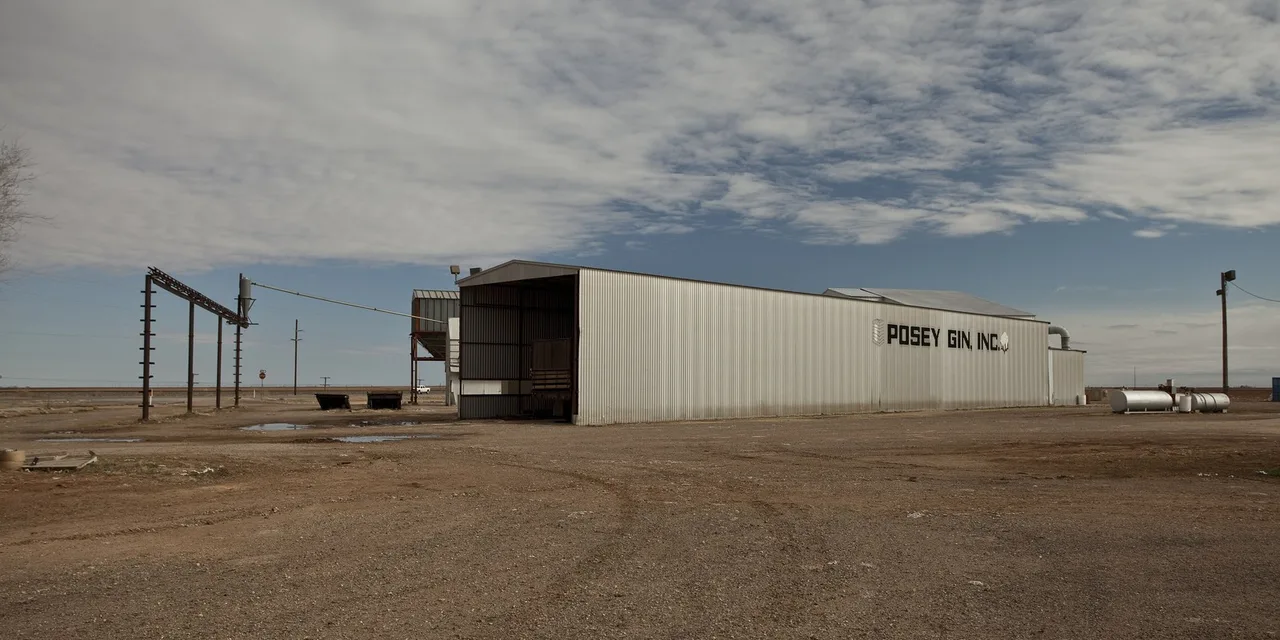 The long metal building of Posey Gin, Inc., a cotton gin at Posey in Lubbock County, with an overhead pipe on posts to the left and tanks to the right, under a wide sky