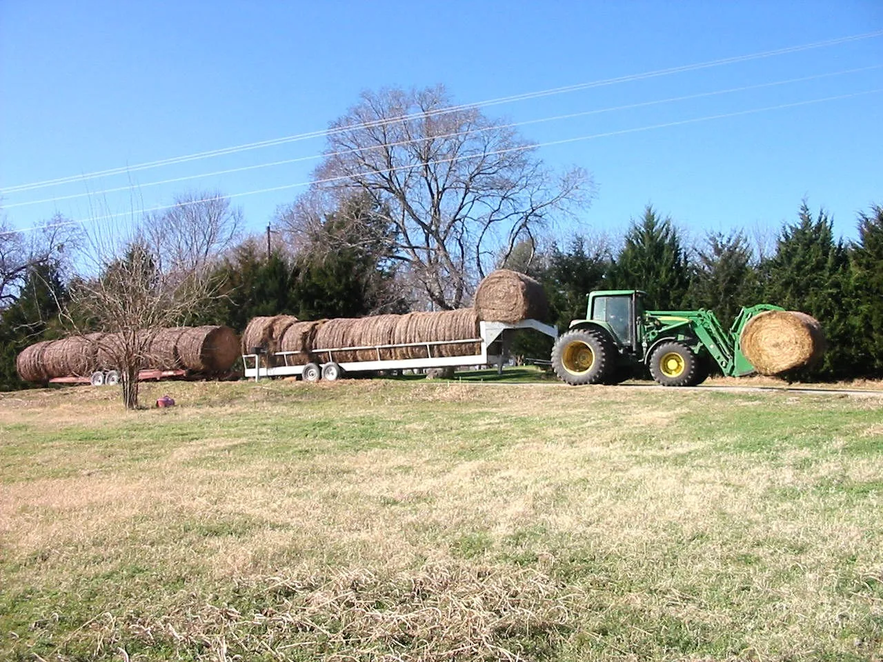 A green tractor with a front loader carrying a round hay bale and pulling two trailers of round bales along a rural road in Grayson County, Texas