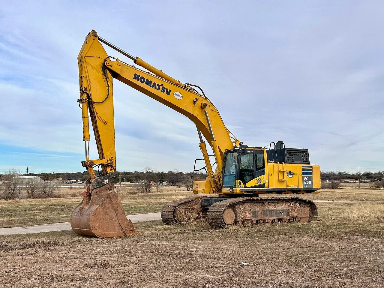 A yellow Komatsu PC650LC crawler excavator parked on dry grass in Tom Green County, Texas, with its bucket resting on the ground and houses in the distance