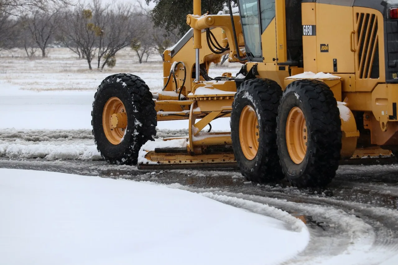A yellow G930 motor grader clearing slush from a curved road at Dyess Air Force Base in Abilene, with one front wheel, the blade and the rear wheels in the frame and snow on the ground behind
