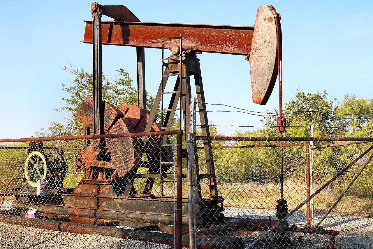 A rust-red oil well pumping unit behind a chain-link fence in Lake Arrowhead State Park near Wichita Falls, with trees behind it