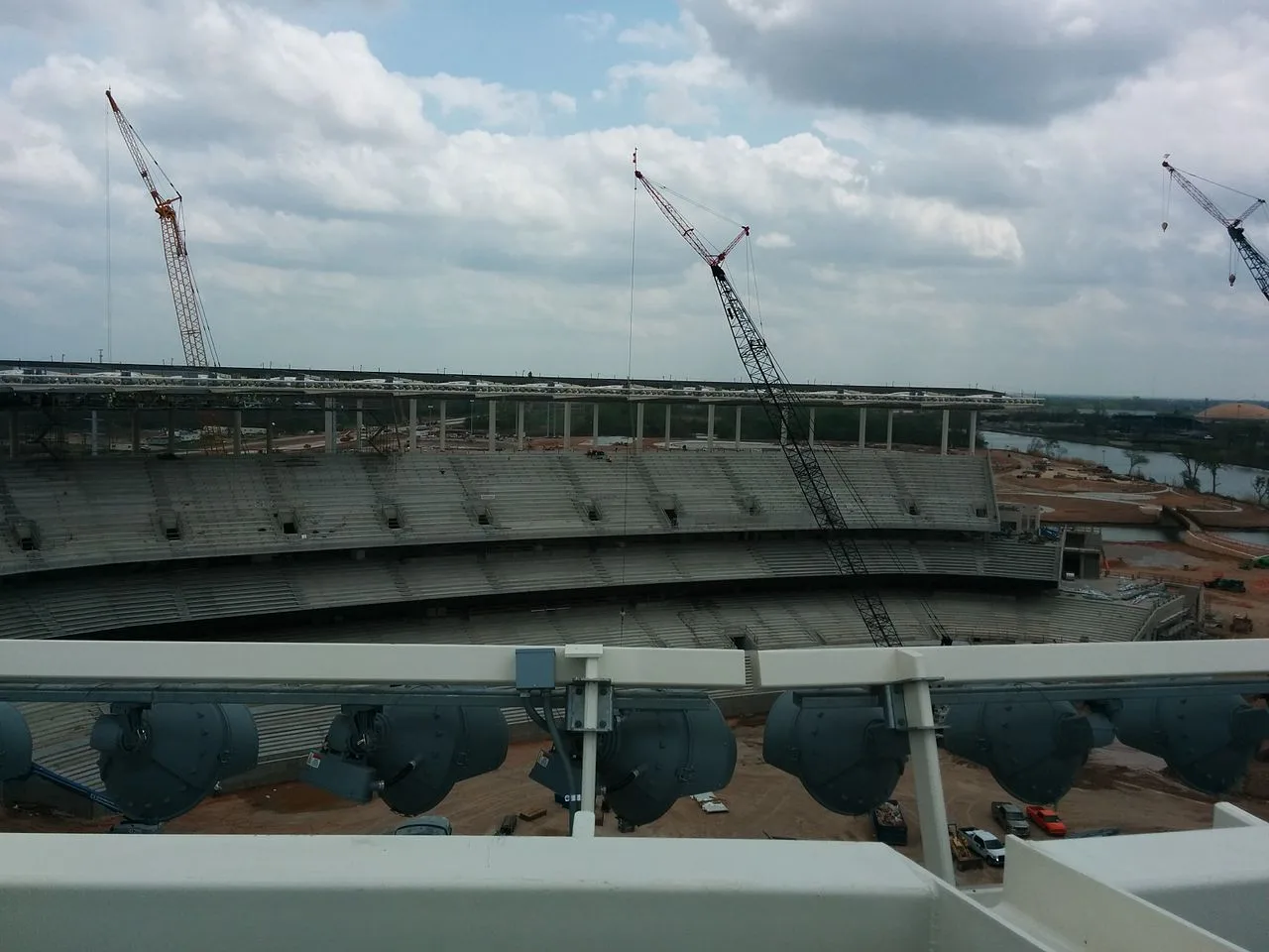 Three lattice boom cranes around the concrete seating bowl of McLane Stadium in Waco while it was under construction, with a river beyond the stands on the right