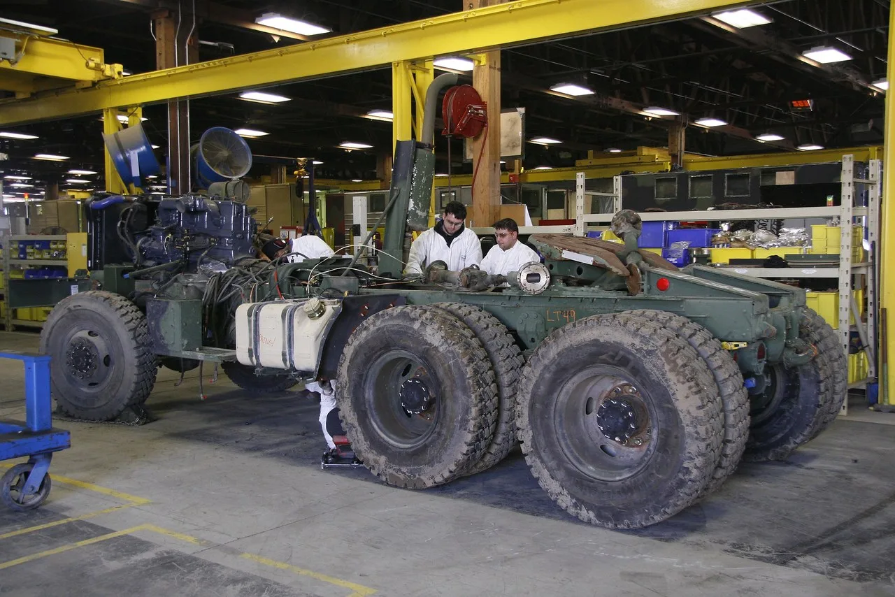 A heavy military truck chassis with its engine exposed on a shop floor under yellow overhead crane beams, with two workers in white coveralls behind it