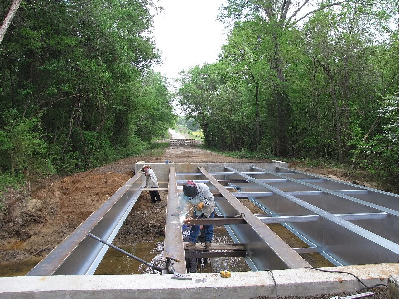 A Smith County Road and Bridge crew at work on a new bridge over a creek on County Road 2125, one man welding between steel girders and a second standing at the left