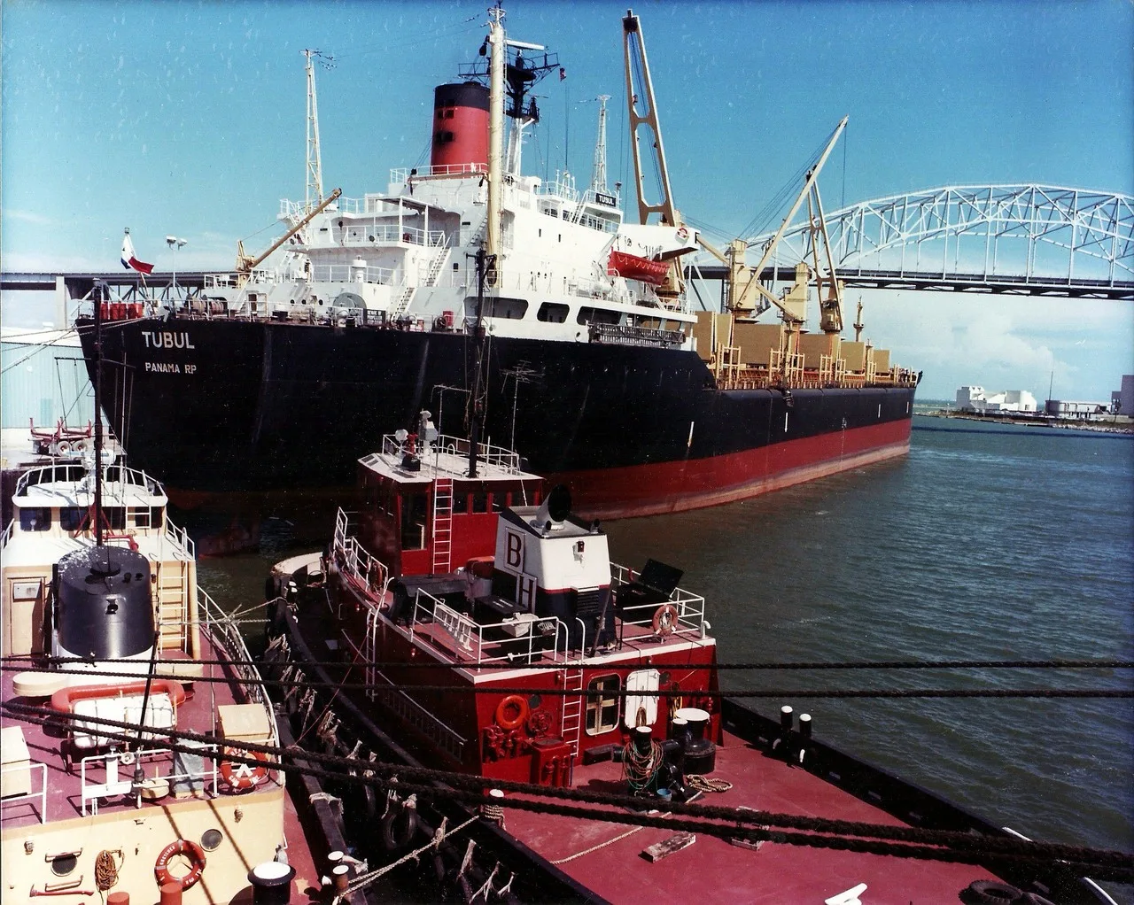 Two tugboats alongside the cargo ship Tubul in the Port of Corpus Christi, with the old Harbor Bridge behind