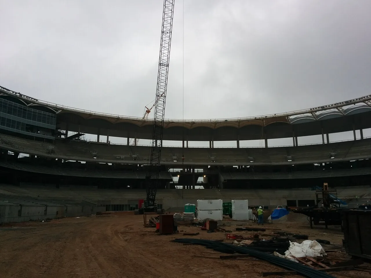 A lattice boom crane standing inside the seating bowl of McLane Stadium in Waco during construction, with building materials and a worker on the dirt floor
