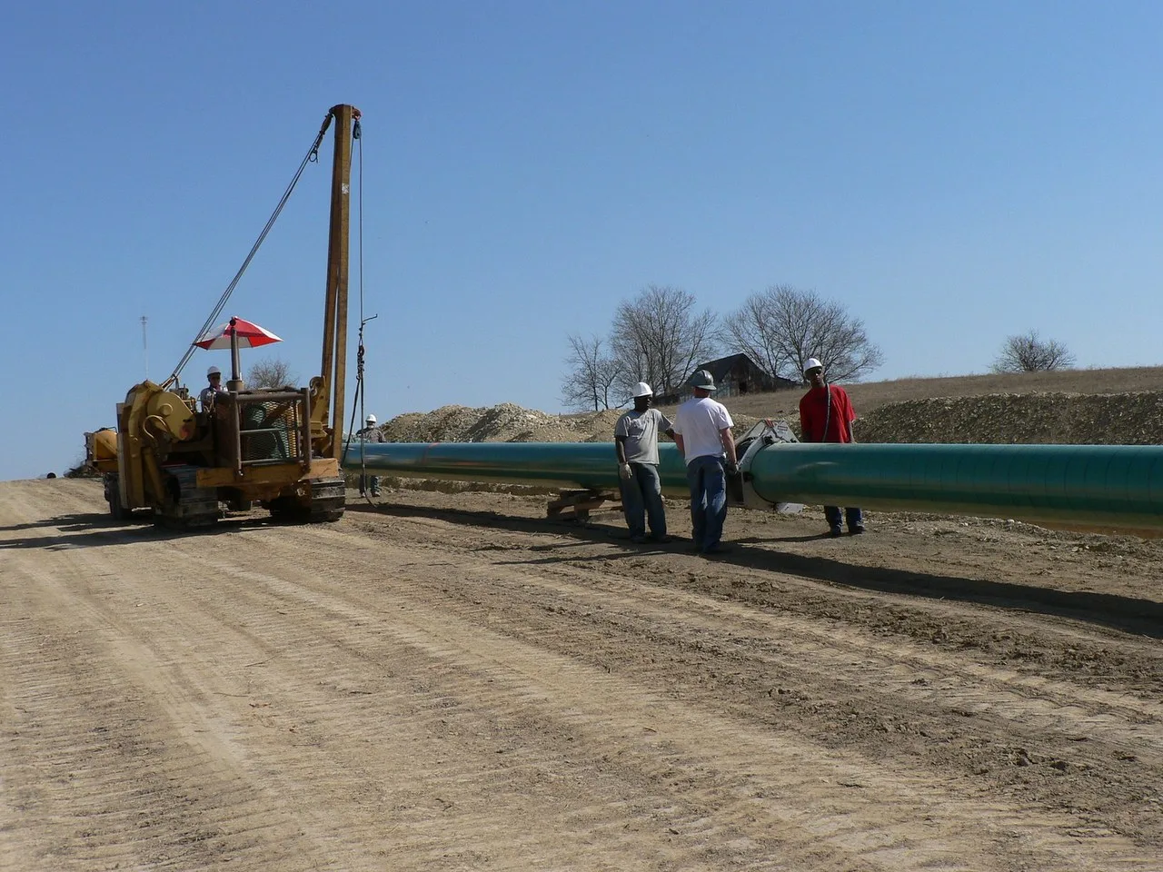 A yellow side boom pipelayer on tracks beside a length of green-coated pipe on a dirt right-of-way in Grayson County, Texas, with three workers in hard hats at the pipe