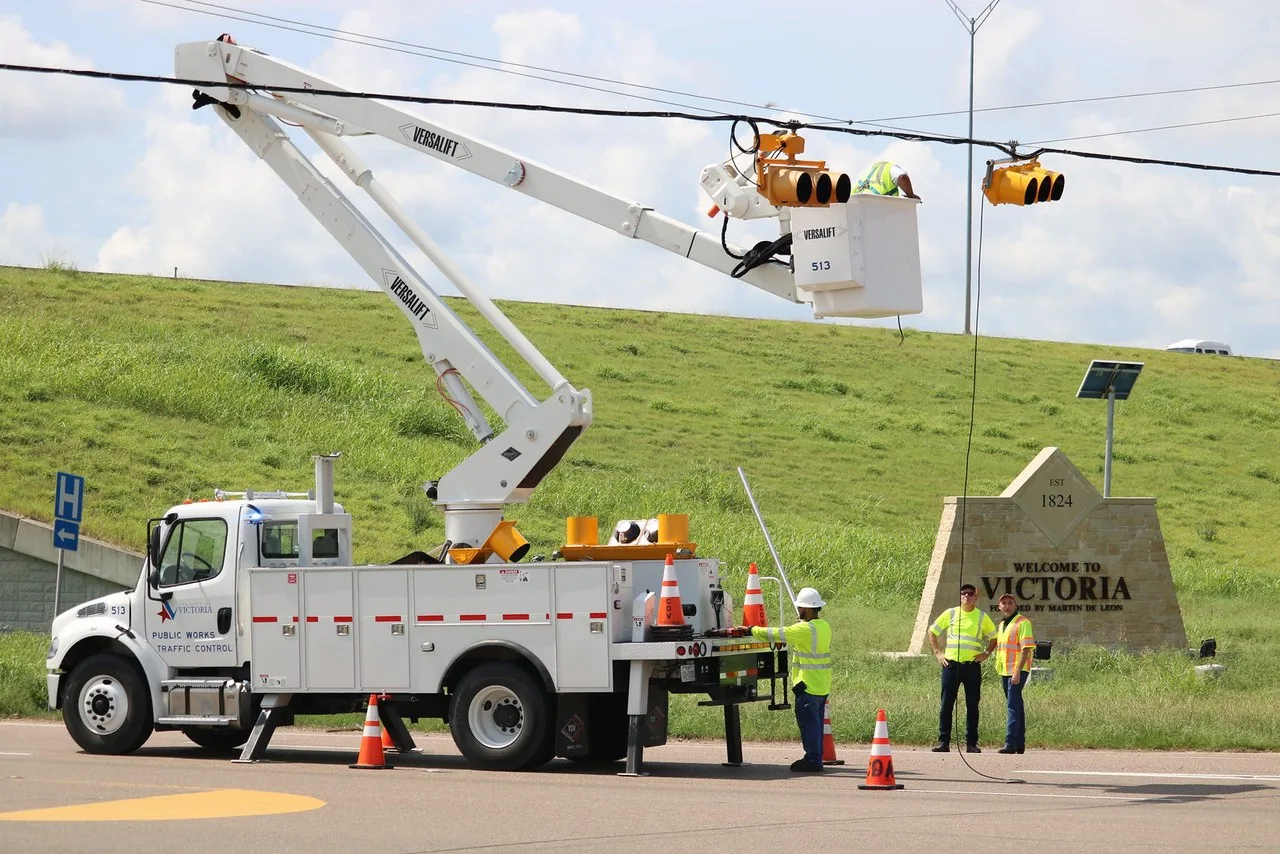 A white Versalift bucket truck marked Victoria Public Works Traffic Control raising a worker to a traffic signal beside a stone Welcome to Victoria sign
