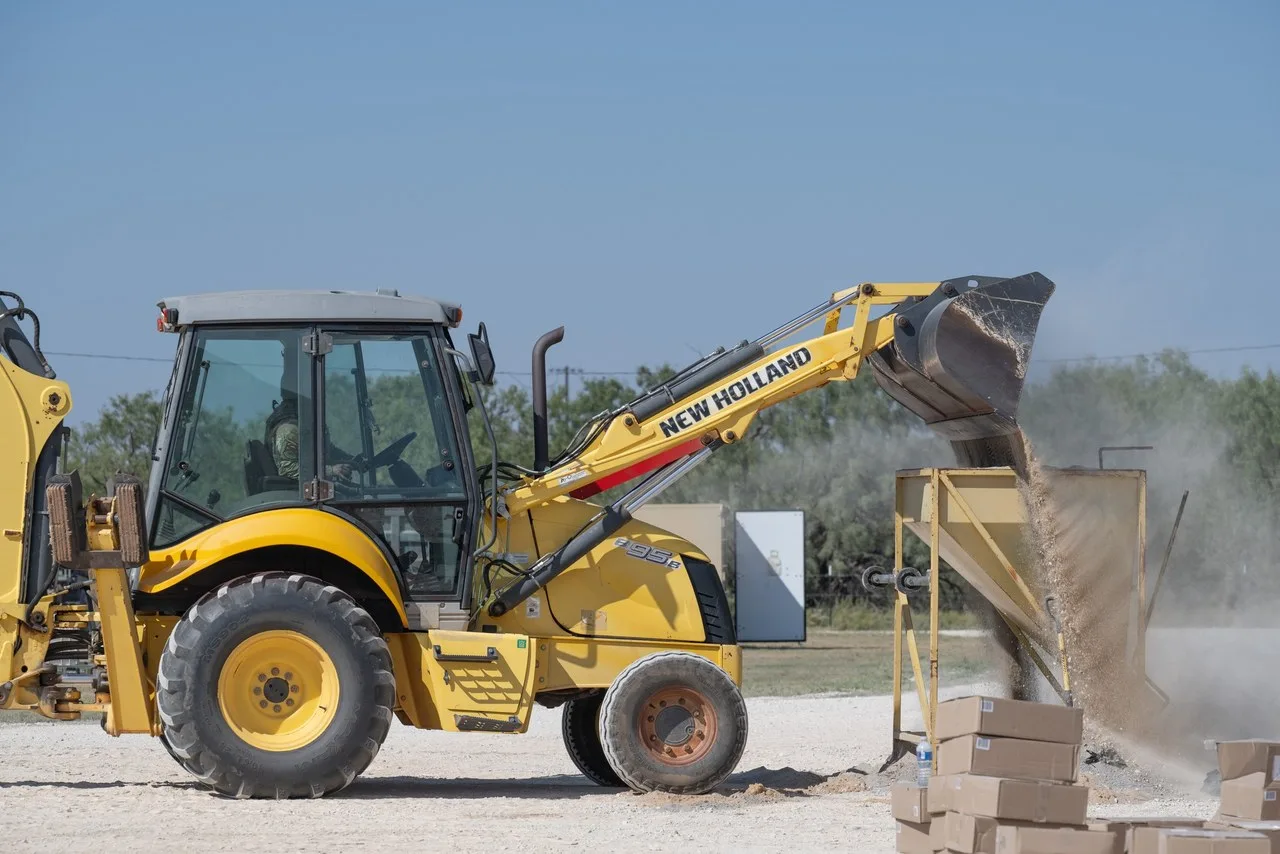 A yellow New Holland B95B backhoe loader tipping its front bucket of loose sand or gravel into a hopper on a gravel yard at Dyess Air Force Base in Abilene, with dust in the air and trees behind