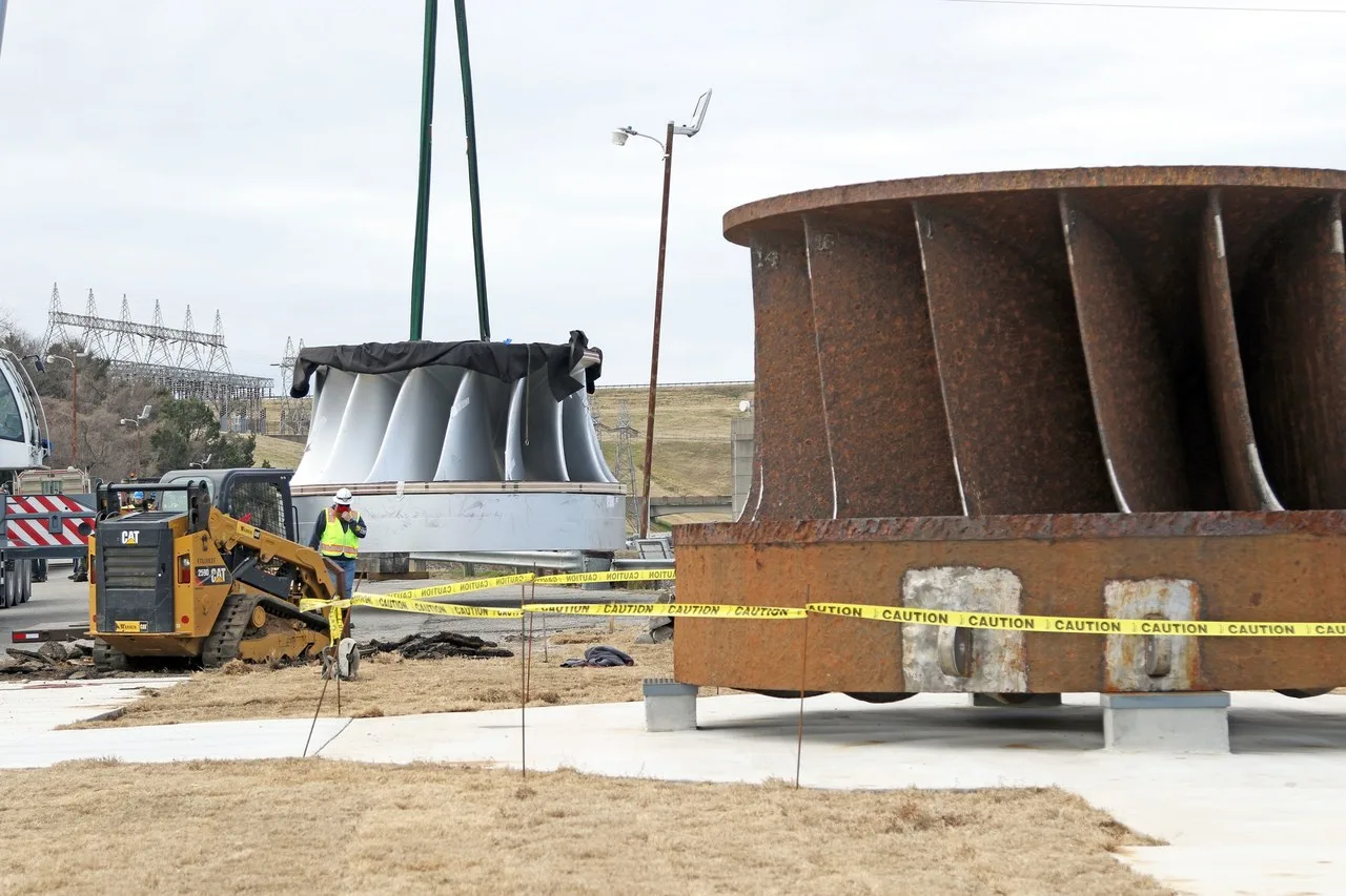 A yellow Cat compact track loader and a worker beside a new turbine wheel hanging from lifting slings, with the rusted old one in the foreground, at Denison Dam on Lake Texoma