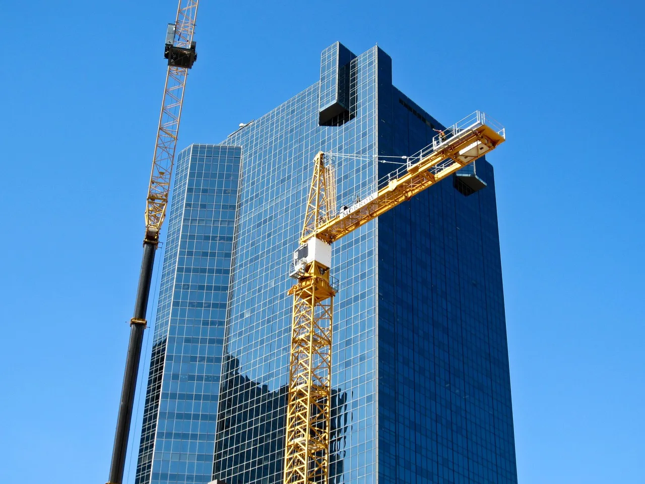A yellow tower crane going up beside a mobile crane in front of a glass office tower in downtown Fort Worth
