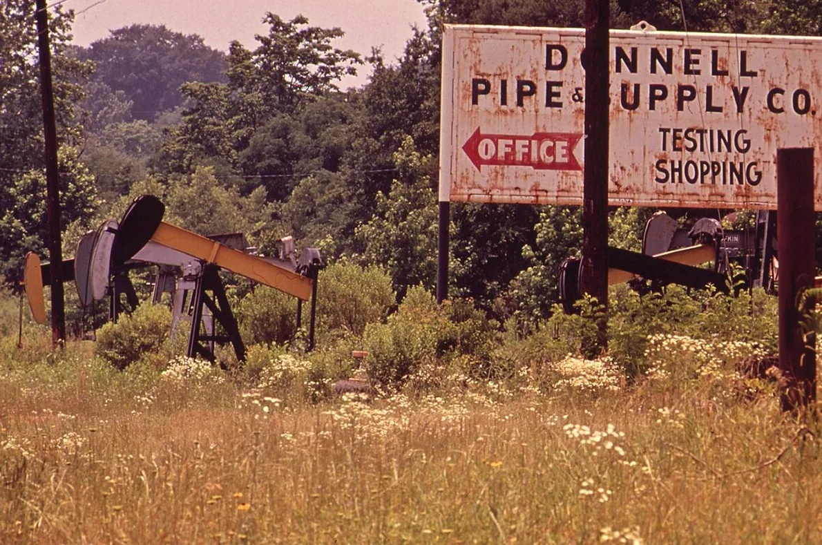 Two oil well pumpjacks behind tall grass and brush beside a rusted pipe and supply company sign in Kilgore, in an old color photograph