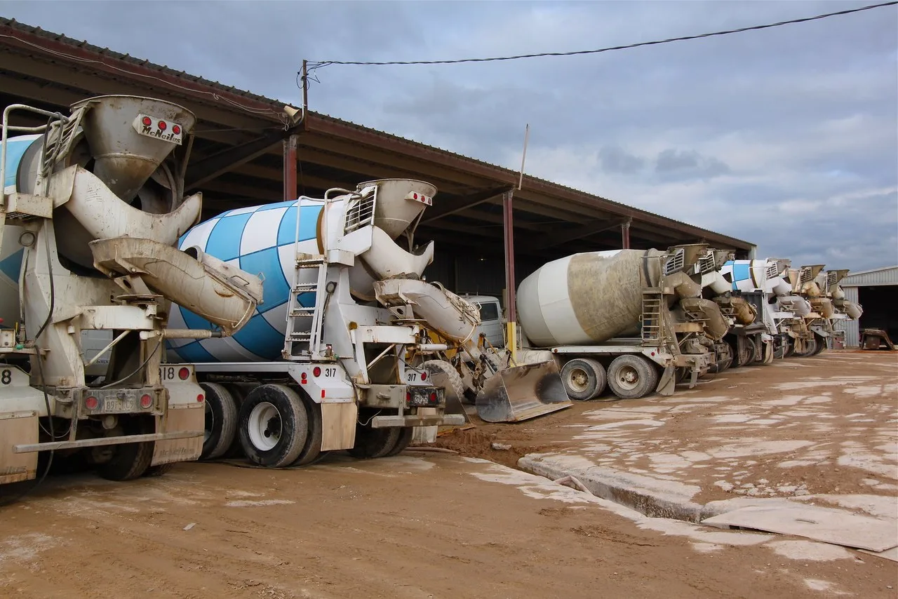 A row of concrete mixer trucks parked beside an open-sided shed at a concrete plant in Wichita Falls, with a loader bucket showing between two of them
