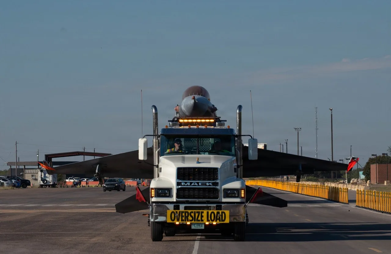 A white Mack semi truck with an Oversize Load sign on its bumper hauling a fighter jet on a trailer along a road at Goodfellow Air Force Base in San Angelo, seen head-on