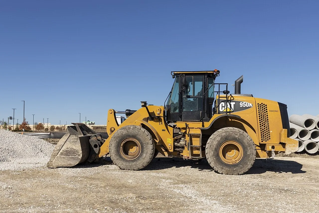 A yellow Caterpillar 950K wheel loader parked on gravel with its bucket on the ground, beside a gravel pile and a stack of concrete pipe