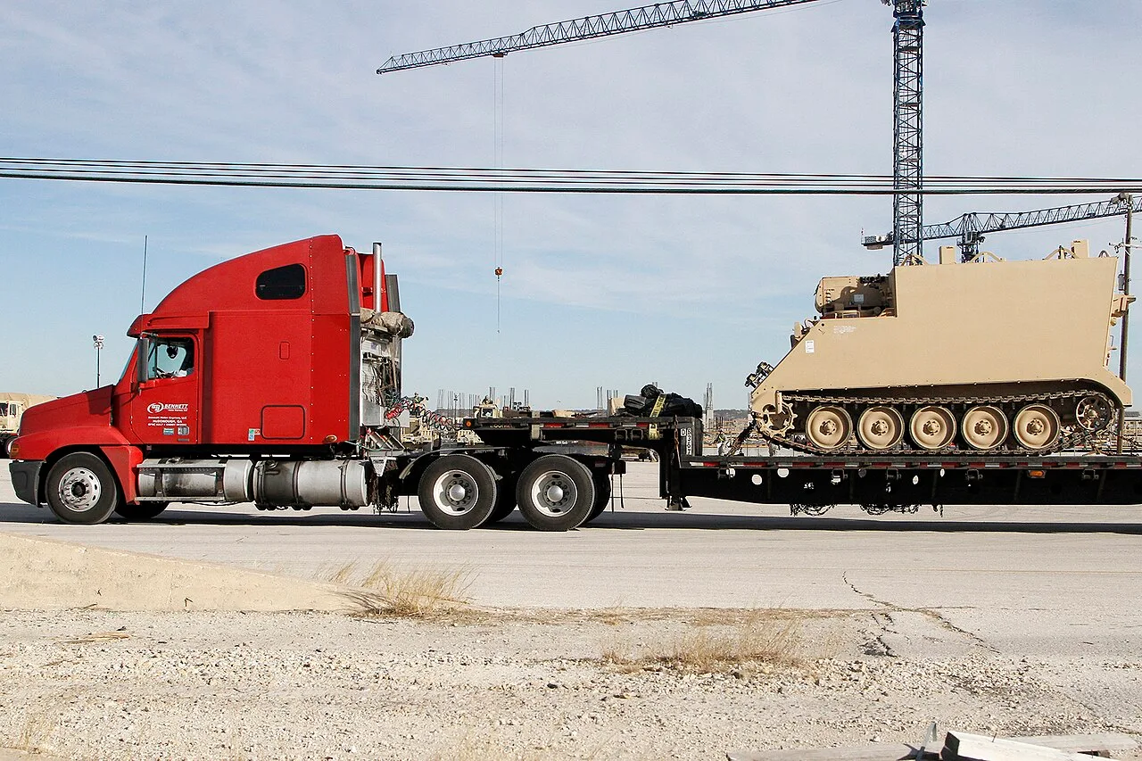 A red semi truck pulling a low trailer loaded with a tracked Army command post vehicle at Fort Hood, with tower cranes in the background