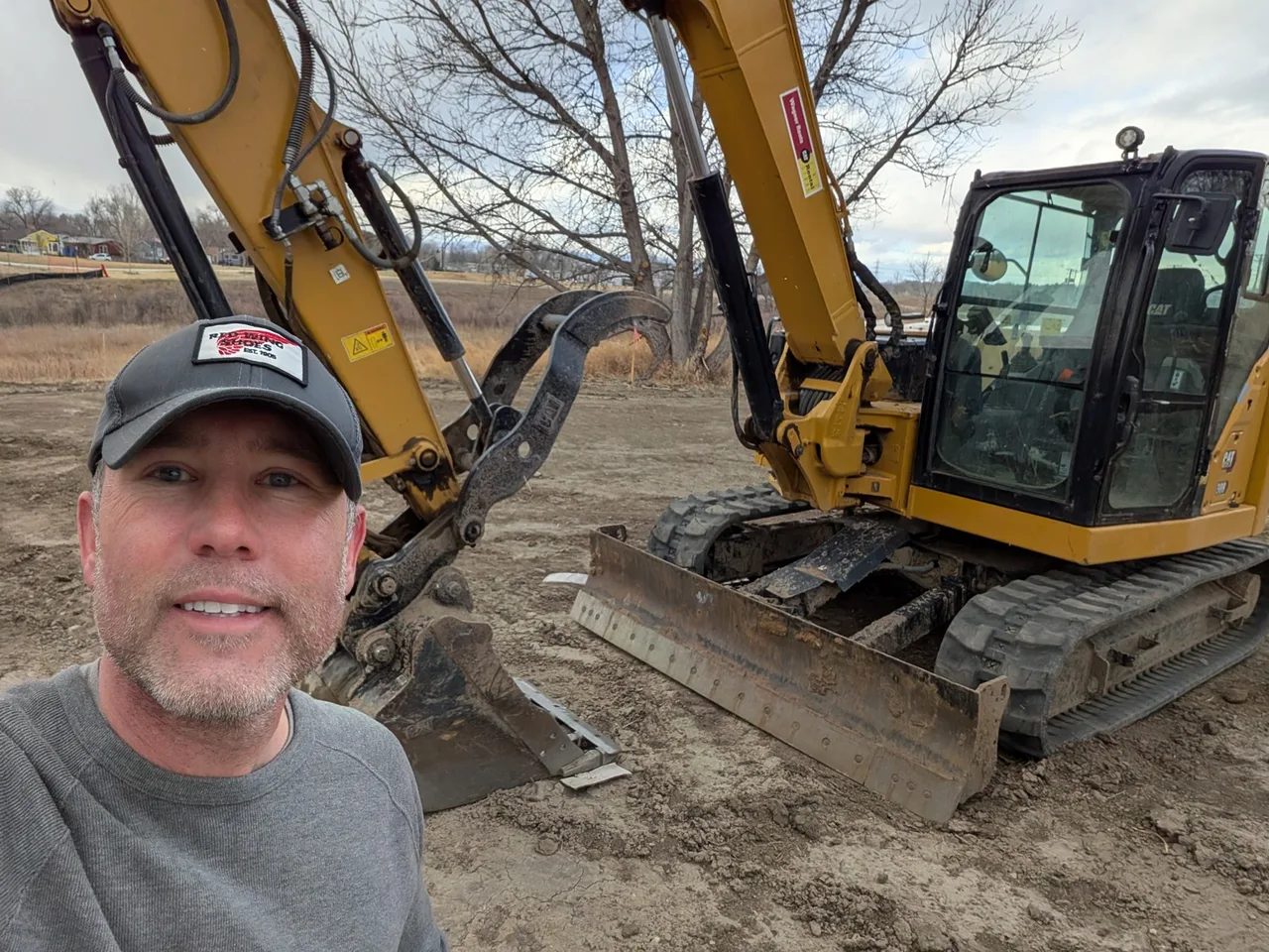 The appraiser in a gray cap beside a yellow Cat compact excavator with a dozer blade and a hydraulic thumb on its bucket.
