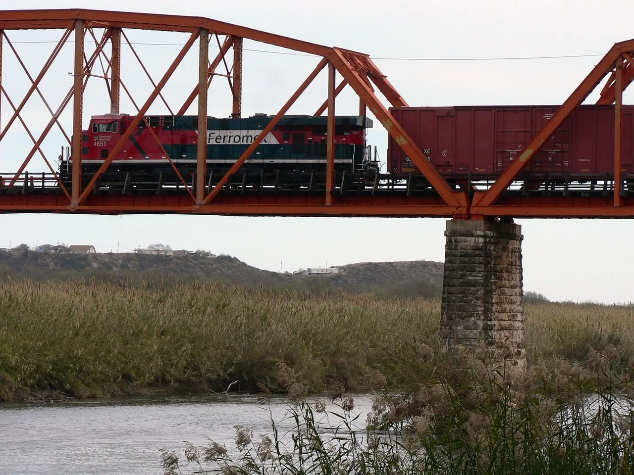 A red and green Ferromex locomotive and a boxcar crossing the orange steel rail bridge between Eagle Pass and Piedras Negras, above the river and its reeds