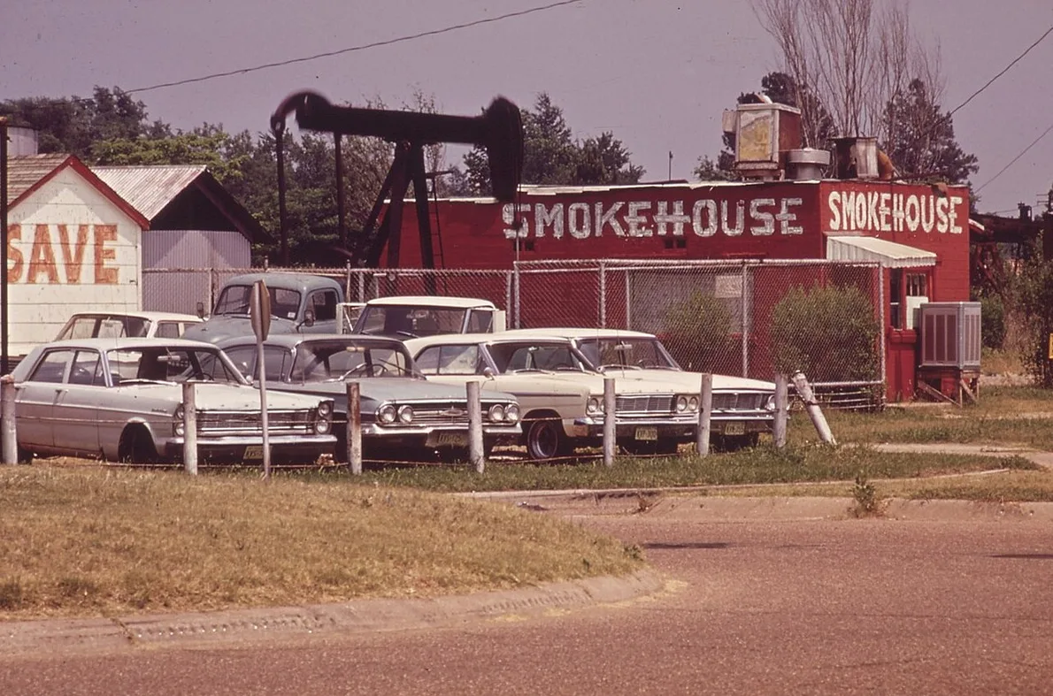 A pumpjack standing behind a red smokehouse restaurant and a row of used cars in Kilgore, in an old color photograph