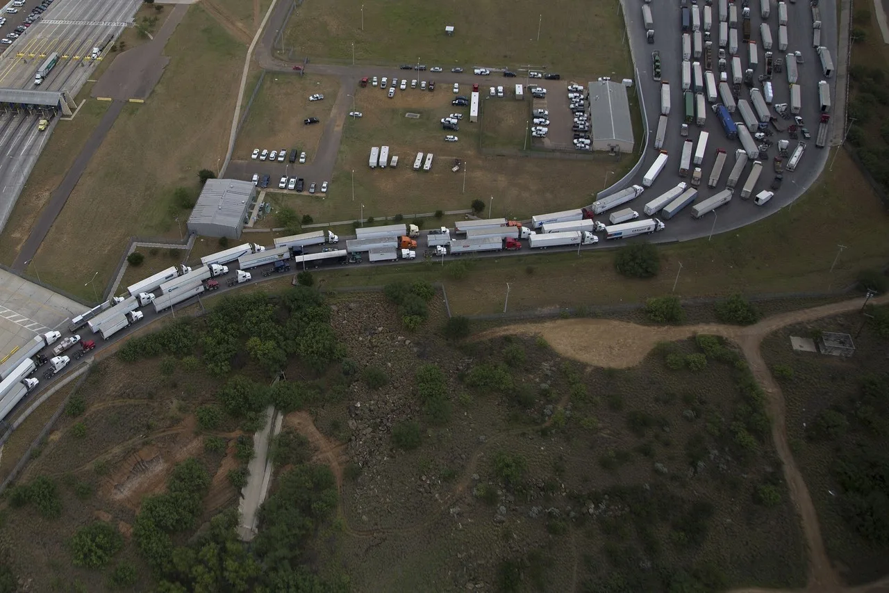 Tractor-trailers lined up nose to tail on a curved road beside a customs facility at the World Trade Bridge in Laredo, seen from the air
