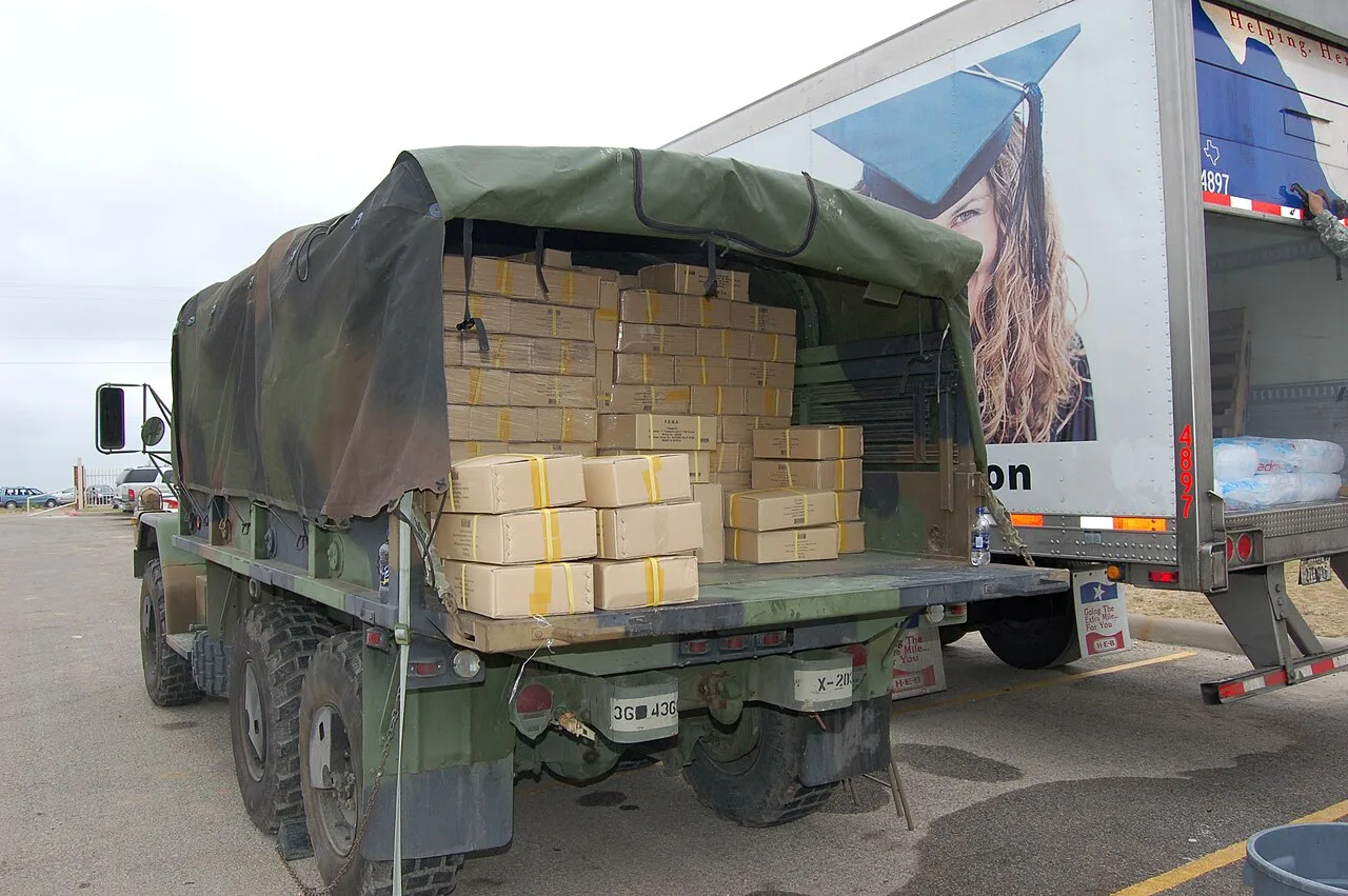 A military cargo truck with a camouflage cover, loaded with taped boxes, parked beside a white box trailer in a parking lot in Eagle Pass, Texas