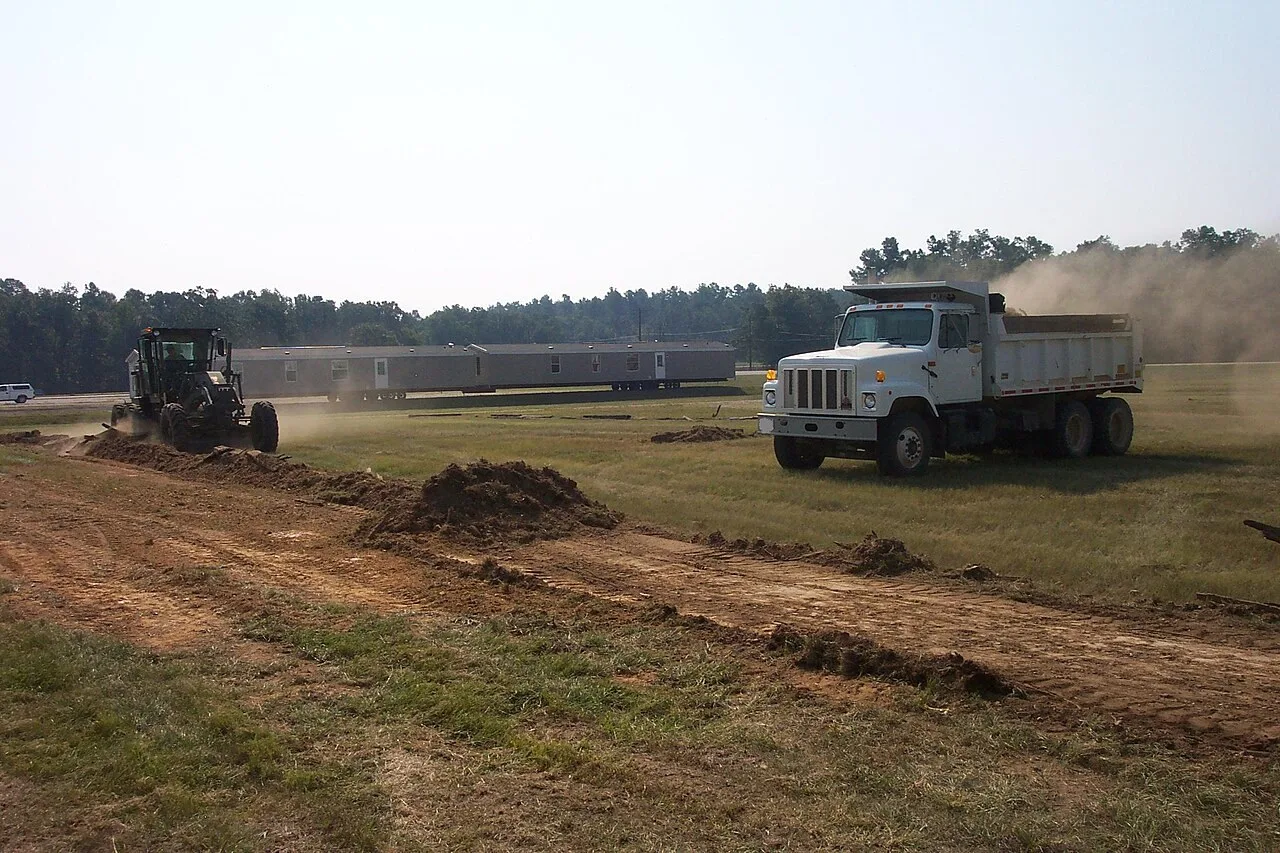 A motor grader and a white tandem dump truck working a strip of dirt in a field at the Lone Star Army Ammunition Plant at Texarkana, Texas, with a long gray mobile home behind them