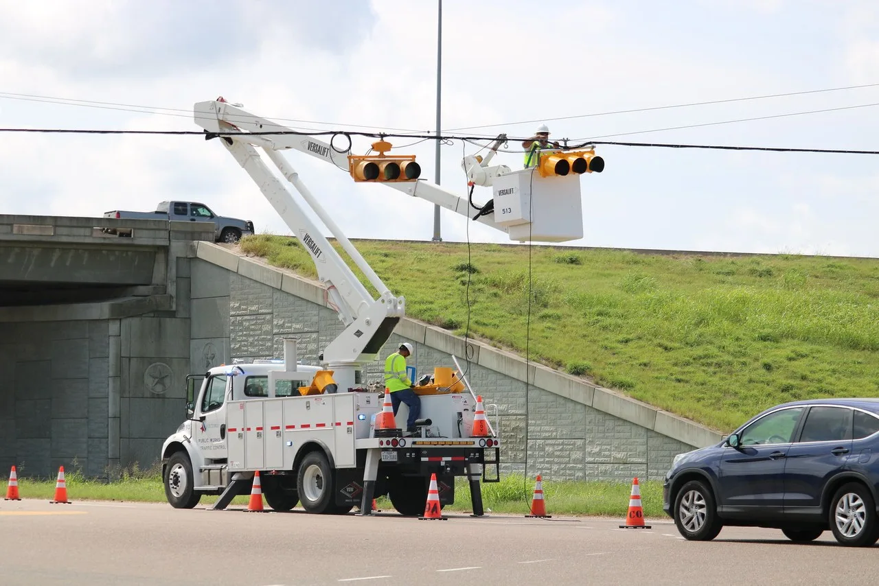 A white bucket truck with its boom raised to traffic signals on a span wire beside an overpass in Victoria, with traffic cones around it