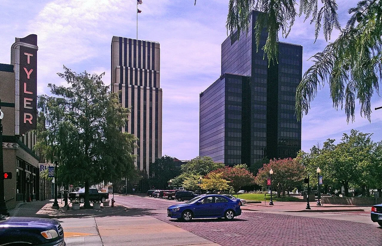 Downtown Tyler seen from a brick street, with a tall vertical TYLER sign on the left and two office towers beyond the trees