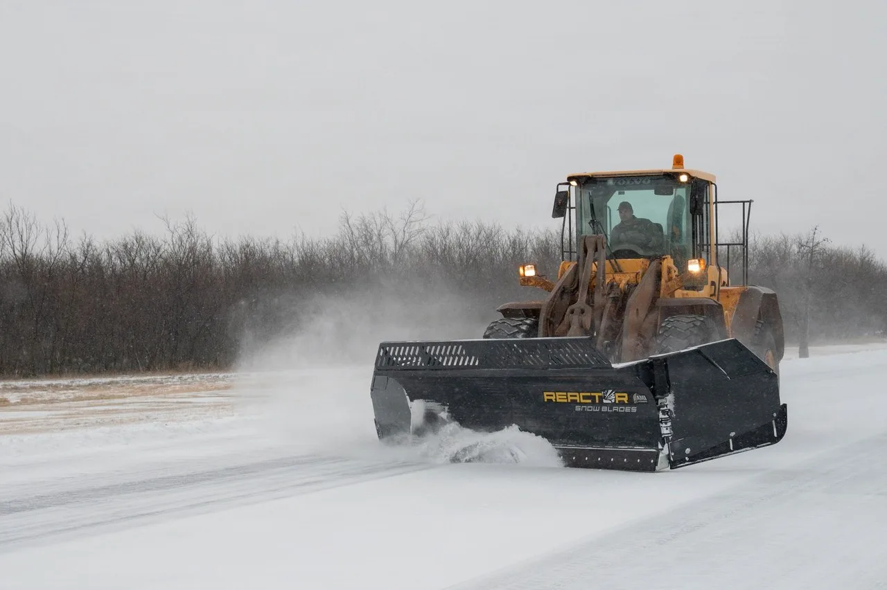 A Volvo wheel loader pushing snow with a wide black snow blade at Dyess Air Force Base in Abilene during a winter storm, with bare trees behind