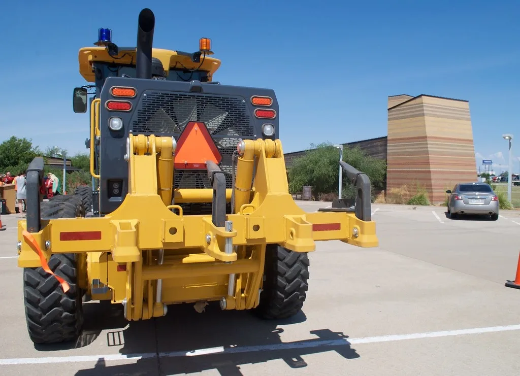 The back of a yellow motor grader parked on a paved lot in Amarillo, with a mounting frame across the rear and an orange slow-moving-vehicle triangle