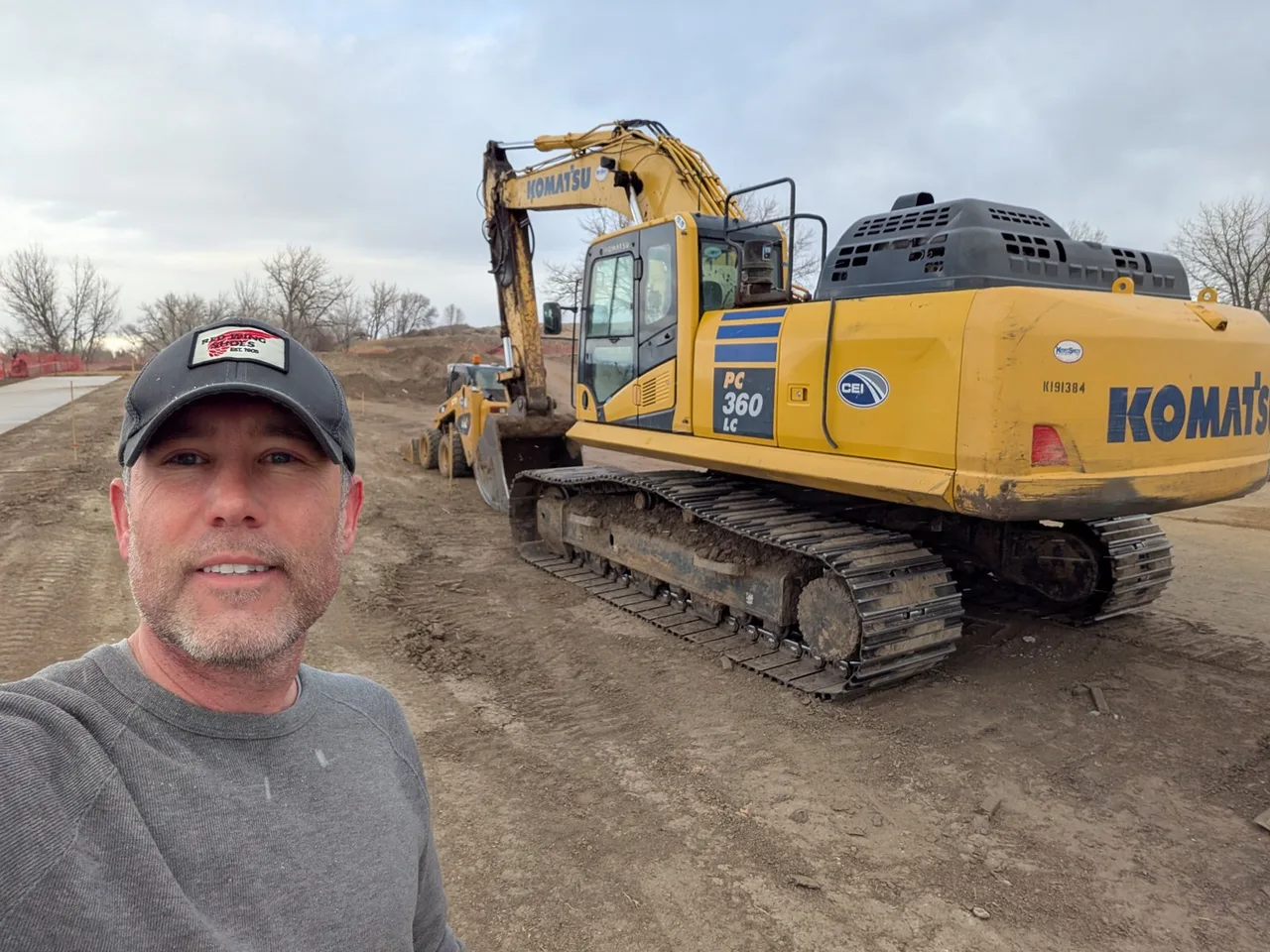 The appraiser in a gray cap on a dirt job site in front of a yellow Komatsu PC360LC crawler excavator.