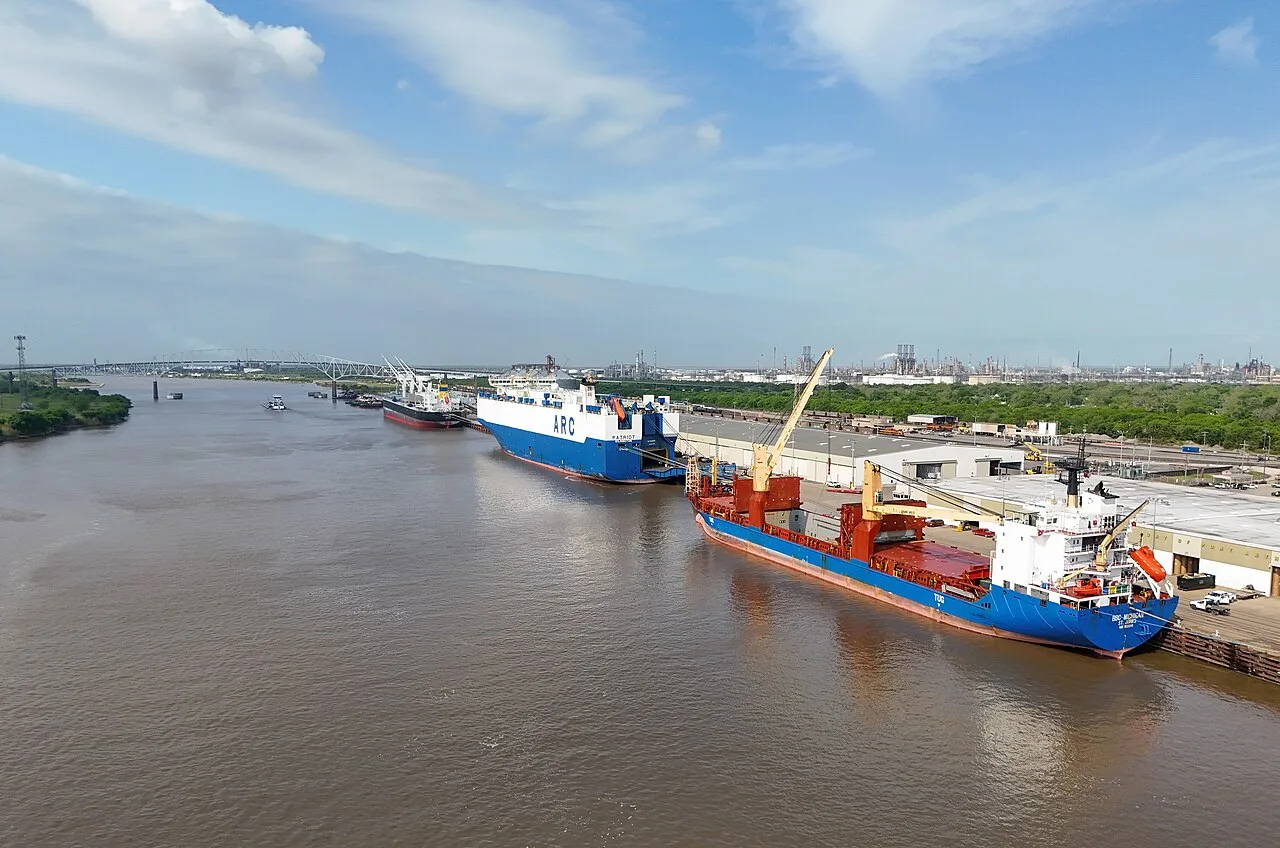 Cargo ships at the Port of Port Arthur wharf seen from above, the nearest one carrying its own deck cranes, with a refinery on the horizon