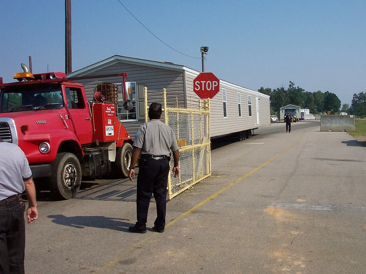A red L9000 truck pulling a mobile home beside a yellow gate and a stop sign at the Lone Star Army Ammunition Plant at Texarkana, Texas, with a man in a gray uniform shirt standing in front of the sign