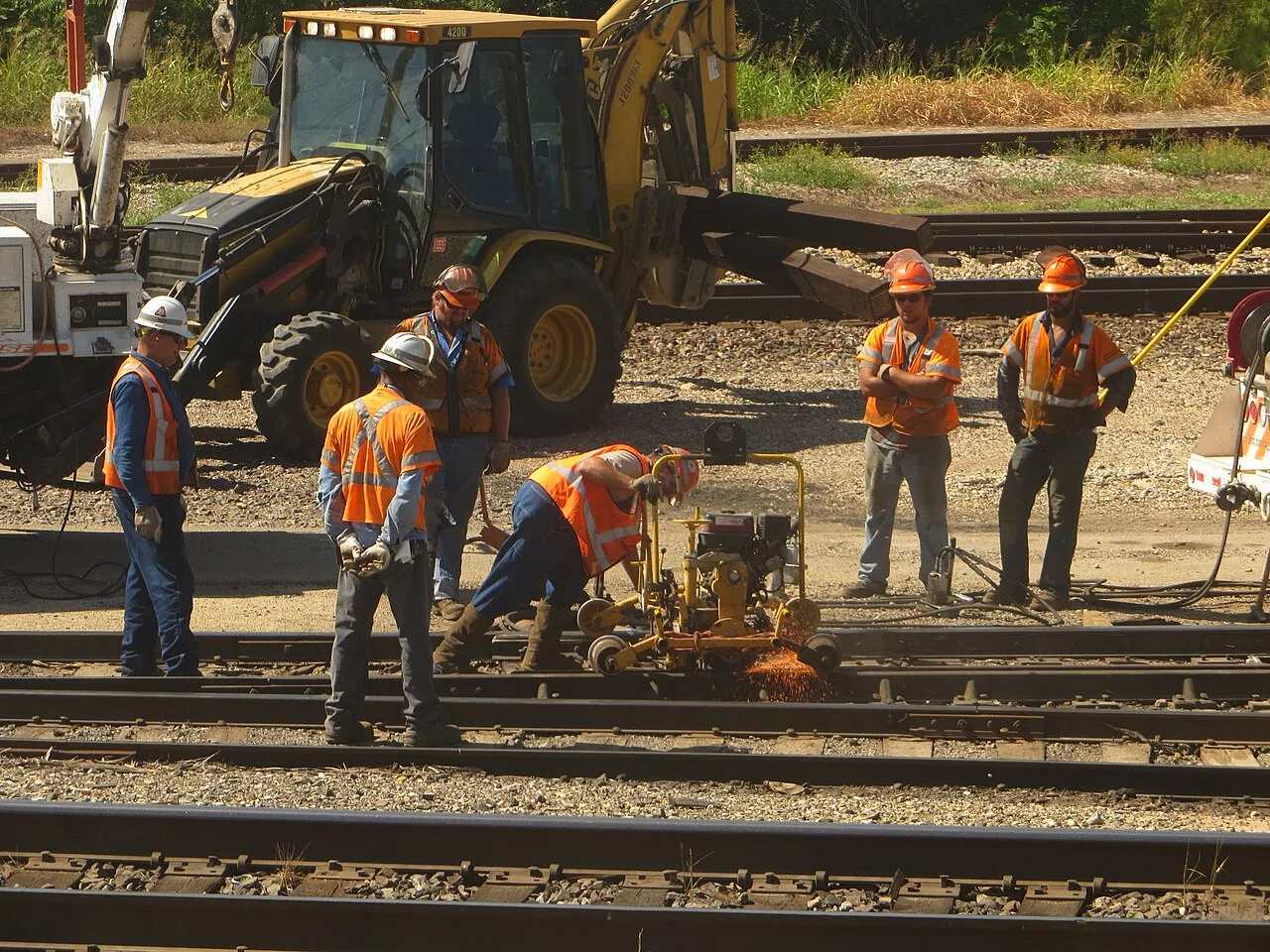 A track crew in orange vests cutting rail beside a Caterpillar backhoe loader carrying crossties on BNSF track in Temple