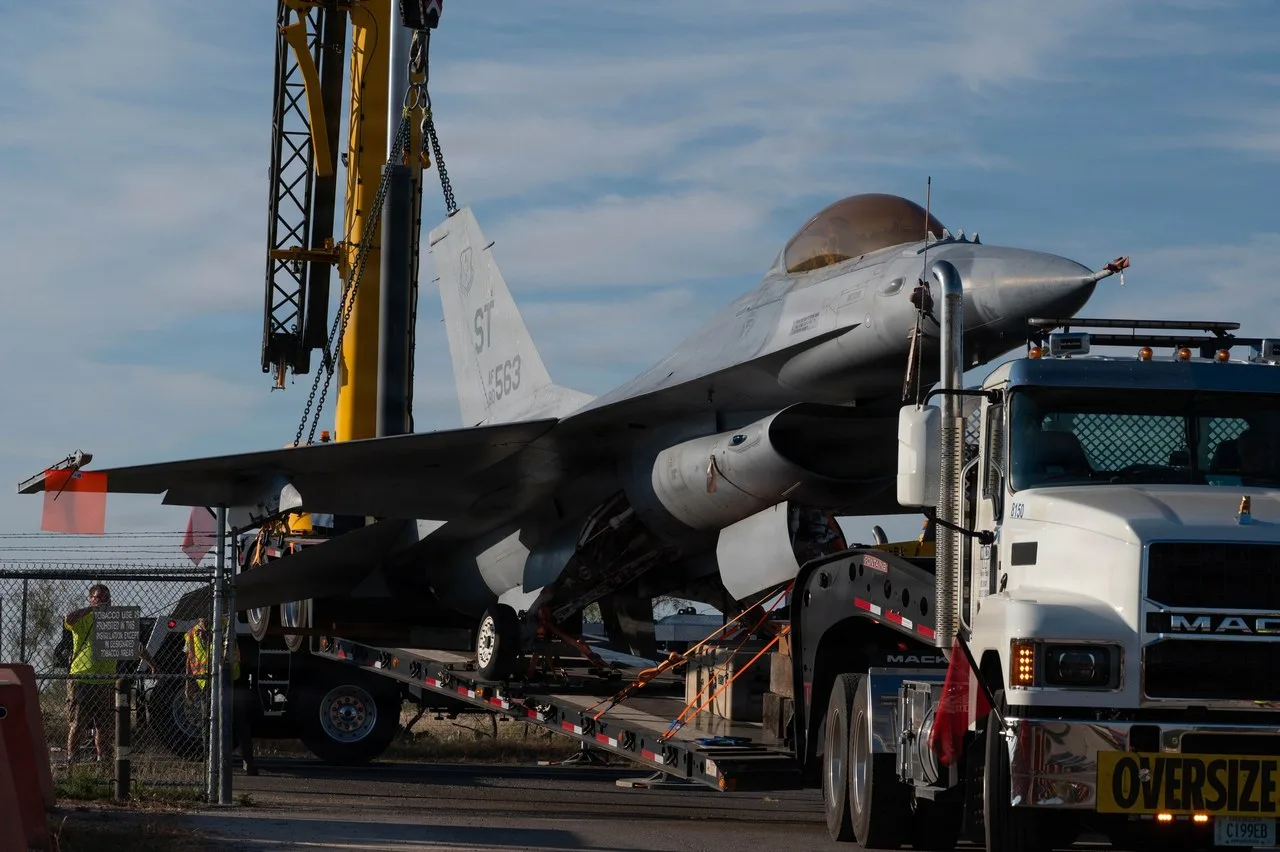 A yellow crane boom with chains above a fighter jet loaded on a low trailer behind a white Mack truck at Goodfellow Air Force Base in San Angelo, with two workers in safety vests at a fence