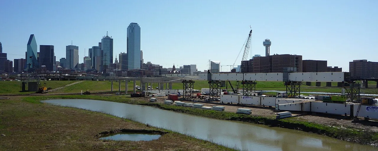 Cranes and steel deck sections during construction of the Margaret Hunt Hill Bridge over the Trinity River, with the downtown Dallas skyline behind